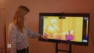 Een foto van een wetenschapper, ze staat bij een scherm. Daarop een beeld van een hittecamera van iemand die fietst. Sommige delen, zoals het hoofd, zijn licht gekleurd en dus erg warm. De benen zijn donkerder gekleurd en dus koeler.