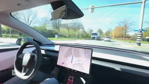 Een foto vanuit een auto. De auto rijdt op de weg, richting een kruising met stoplichten. Er zit een bestuurder, maar die houdt zijn handen niet aan het stuur. Op het dashboard staat een groot display met een plattegrond.