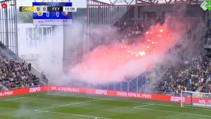 Foto van voetbalstadion waar oranje fakkels worden afgestoken op de tribune, wat grote rookwolken veroorzaakt.