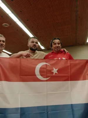 Een foto van drie mannen in een gymzaal. Ze houden een rood-wit-blauwe Nederlandse vlag vast met daarin de Turkse vlag verwerkt, een witte maan en ster op een rood vlak.