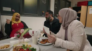 Een foto van twee vrouwen met een hoofddoek en presentator Hasan aan een tafel in een buurthuis. Op de tafel staan snacks zoals churros en snackgroente.
