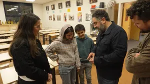 Een foto van presentator Hasan met drie leerlingen in een klas. Hasan wijst op een foto die hij in zijn hand heeft, de leerlingen kijken ernaar.