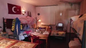 Een foto van een gezellig kamertje met stapelbedden tegen de muur. Er staat een tafel met stoelen in het midden, er staat een kleine tv en er hangt een Turkse vlag aan de muur.