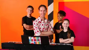 Foto van zangcoach Michiel met kinderen van Kinderen voor Kinderen in zwarte t-shirts voor kleurrijk decor en bij piano.