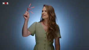 Een foto van de presentatrice in de studio. Ze houdt een model van een clitoris omhoog. Het is een klein V-vormig ding van roze plastic dat bijna in een handpalm past.