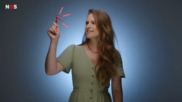 Een foto van de presentatrice in de studio. Ze houdt een model van een clitoris omhoog. Het is een klein V-vormig ding van roze plastic dat bijna in een handpalm past.