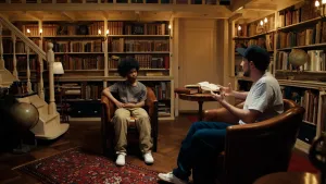 Foto van jongen met afrokapsel in gesprek met jonge man met pet, beide in bruine leren stoelen voor oude boekenkasten met sfeerverlichting.