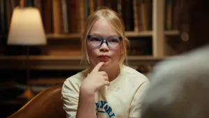 Foto van blond meisje met blauwe bril voor een oude boekenkast. Ze houdt haar wijsvinger aandachtig luisterend tegen haar kin.